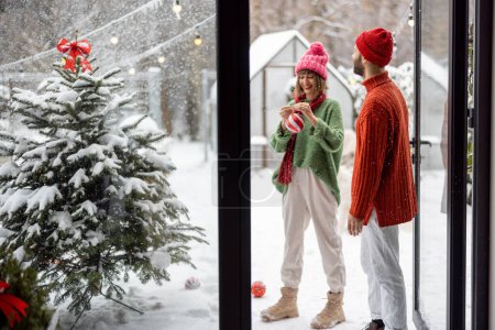 Foto de Man and woman prepare to decorate Christmas tree with festive balls, standing together happily at snowy backyard. Young family celebrating winter holidays at home - Imagen libre de derechos