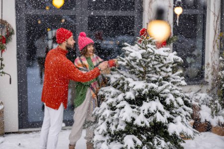 Foto de Man and woman decorate Christmas tree with festive balls, while preparing for a winter holidays at snowy terrace of their house. Image focused on tree, people on background - Imagen libre de derechos