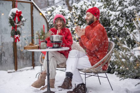 Foto de Man and woman have romantic dinner with fondue, while sitting together by the table at beautifully decorated snowy backyard. Young family celebrating winter holidays outdoors - Imagen libre de derechos