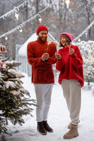 Foto de Man and woman in red sweaters celebrate New Years holidays by lighting sparklers and having fun near Christmas tree at snowy backyard - Imagen libre de derechos
