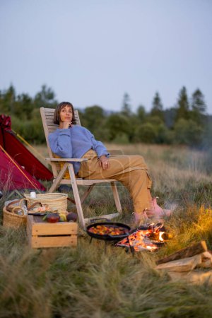 Foto de Mujer tiene un picnic, sentado junto a la hoguera en la silla de madera cerca de la tienda de campaña, viajando en las montañas en la hora de verano - Imagen libre de derechos