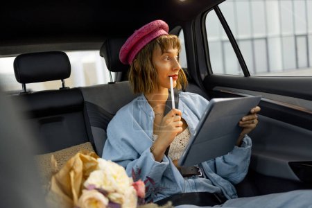 Photo for Young stylish woman uses touchpad, doing some creative work while sitting on backseat at car on the go. Concept of transportation and modern digital lifestyle - Royalty Free Image