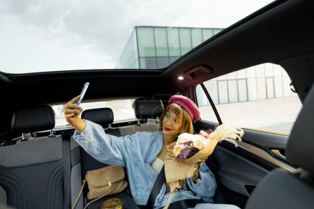 Téléchargez les photos : Jeune femme élégante fait selfie au téléphone ou a un appel vidéo, tout en étant assis avec des fleurs sur le siège arrière d'une voiture. Femme voyageant en voiture à la ville. Vue large, véhicule avec toit panoramique - en image libre de droit