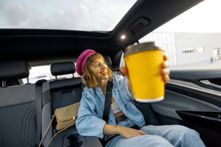 Photo for Stylish woman travels with coffee on backseat of a car, showing yellow coffee cup with blank space. Wide angle view, modern car with panoramic rooftop - Royalty Free Image