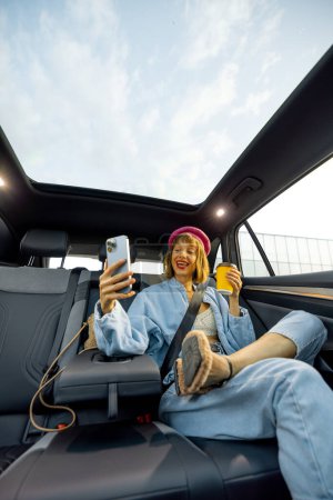 Téléchargez les photos : Jeune femme élégante utilise un téléphone intelligent tout en étant assis détendu avec une tasse de café sur le siège arrière de la voiture sur la route. Vue grand angle, voiture moderne avec toit panoramique - en image libre de droit