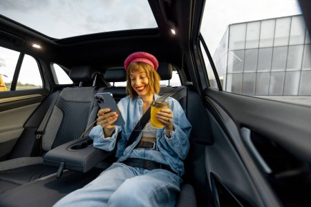 Foto de Mujer joven y elegante utiliza el teléfono inteligente mientras está sentado relajado con una taza de café en el asiento trasero del coche sobre la marcha. Amplia vista angular, coche moderno con techo panorámico - Imagen libre de derechos