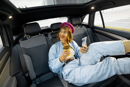 Foto de Joven mujer con estilo disfruta de viajar en coche, sentado relajado con una taza de café y teléfono en el asiento trasero. Amplia vista angular, coche moderno con techo panorámico - Imagen libre de derechos