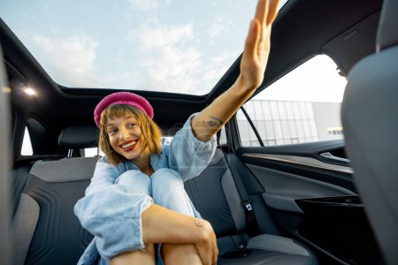 Téléchargez les photos : Jeune femme élégante assise détendue sur le siège arrière d'une voiture moderne tout en voyageant, appelant chauffeur avec une main - en image libre de droit