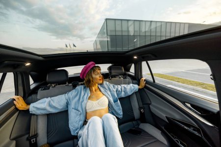 Foto de Mujer joven y elegante sentado relajado en el asiento trasero de un coche moderno mientras viaja por la ciudad. Coche moderno con techo panorámico - Imagen libre de derechos