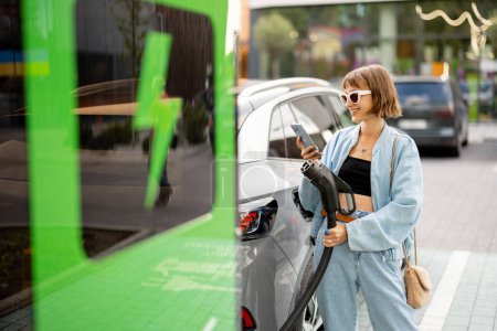 Photo for Young woman with phone and plug for vehicle fast charging standing on a public charging station outdoors. Concept o felectric car charging, electricity and green energy for driving - Royalty Free Image