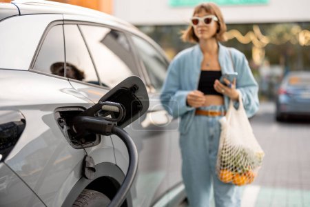 Cloae-up of fast charger plug in vehicle charging port, woman with groceries waiting for her car to be charged on background