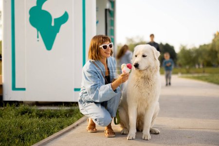 Foto de Mujer alimenta a su lindo perro con un helado mientras caminan juntos en un parque en un día de verano. Amistad con las mascotas y pasar tiempo libre juntos - Imagen libre de derechos
