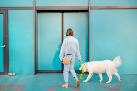 Foto de Mujer con perro blanco que entra en el supermercado con hermosas puertas correderas de color turquesa. Concepto de instituciones públicas o tiendas que admiten mascotas - Imagen libre de derechos