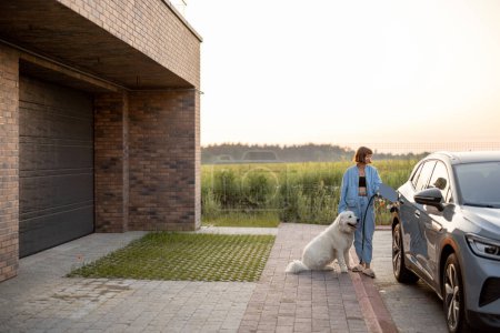 Téléchargez les photos : Femme charge voiture électrique, debout avec son chien blanc mignon près de sa maison de luxe au coucher du soleil. Concept d'énergie verte, durabilité et mode de vie moderne - en image libre de droit