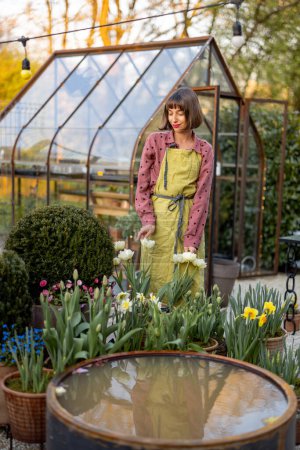 Foto de Retrato de una mujer joven como florista de pie en un hermoso jardín con flores en maceta, plantas verdes y invernadero vintage en el fondo. Concepto de hobby y jardinería - Imagen libre de derechos