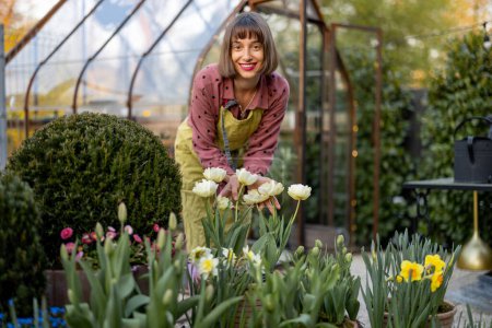 Foto de Retrato de una mujer joven como florista de pie en un hermoso jardín con flores en maceta, plantas verdes y invernadero vintage en el fondo. Concepto de hobby y jardinería - Imagen libre de derechos