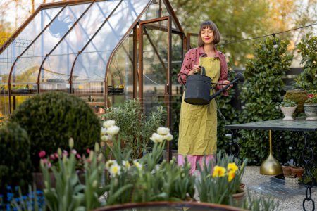 Téléchargez les photos : Portrait d'une jeune femme fleuriste prenant soin des fleurs et des plantes dans le jardin avec serre vintage sur fond. Concept de passe-temps et de jardinage - en image libre de droit