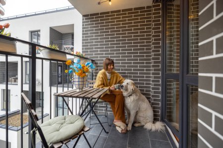 Téléchargez les photos : La jeune femme s'assoit avec son chien sur le balcon, dégustant une boisson au café dans sa propriété pendant la matinée. La vie urbaine dans un complexe résidentiel moderne - en image libre de droit
