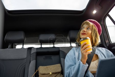 Foto de Mujer joven y elegante con tableta digital y taza de café sentado en el asiento trasero de un coche mientras conduce en la ciudad - Imagen libre de derechos