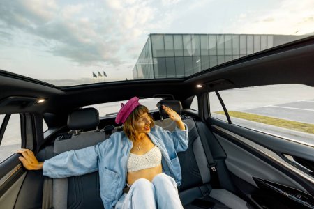 Foto de Mujer joven y elegante sentado relajado en el asiento trasero de un coche moderno mientras viaja por la ciudad. Coche moderno con techo panorámico - Imagen libre de derechos