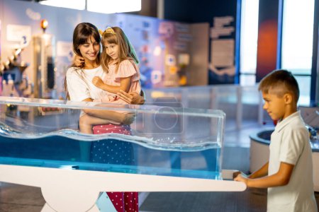 Foto de Mamá con los niños aprenden física interactivamente en un modelo que muestra fenómenos físicos mientras visitan un museo de ciencias. Concepto de entretenimiento y aprendizaje infantil - Imagen libre de derechos