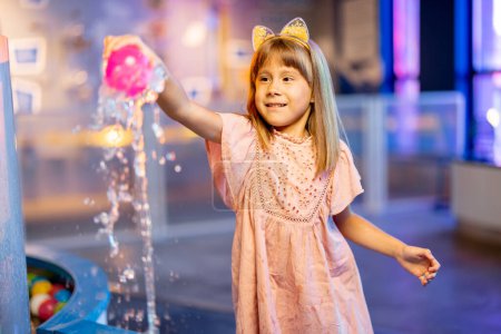Little girl plays with a ball on a steam of water, learning physical phenomena in an interesting way, having fun in a science museum with interactive models