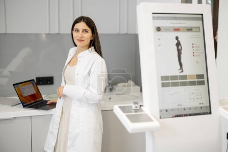 Photo for Young doctor works on laptop while standing near body analyzer machine at beauty salon. Concept of modern technologies in medicine and beauty industry - Royalty Free Image