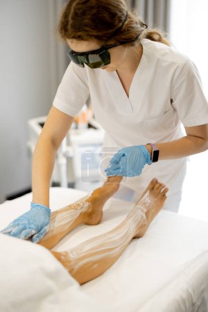 Foto de Trabajador joven de un salón de belleza durante el procedimiento de depilación en las piernas de una mujer. Concepto de depilación y procedimientos de belleza - Imagen libre de derechos
