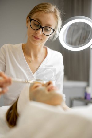Téléchargez les photos : Cosmetologue applique un masque de beauté avec une brosse à un visage de femme dans un salon de beauté. Concept de soins du visage et procédures beauté - en image libre de droit