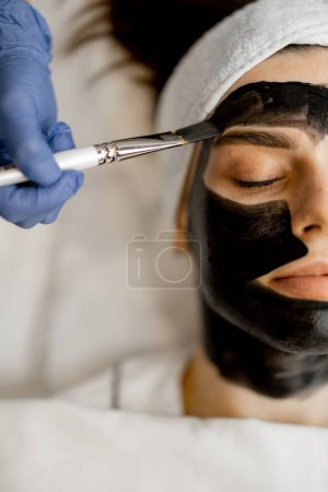 Téléchargez les photos : Appliquer un masque au carbone avec une brosse sur un visage de femme, préparer un peeling au carbone laser dans un salon de beauté, gros plan. Concept de soins du visage et procédures beauté - en image libre de droit