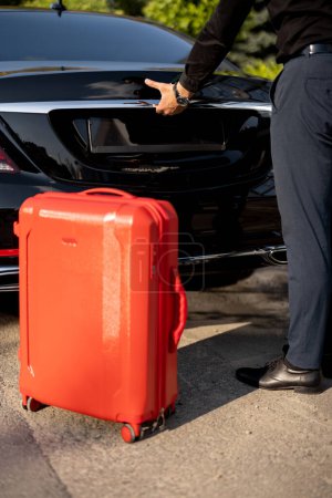 Téléchargez les photos : Le chauffeur met une valise rouge dans un coffre de voiture, gros plan. Concept de voyages d'affaires, chauffeur personnel ou taxi de luxe - en image libre de droit