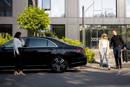 Photo for Man helps a businesswoman to carry her suitcase into a luxury car from an office building or hotel. Female chauffeur waits near a car. Concept of business trips, and luxury transfer services - Royalty Free Image