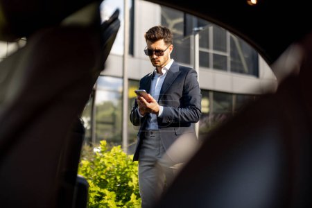 Téléchargez les photos : Beau homme d'affaires parle par téléphone tout en se tenant près de voiture de luxe près de l'immeuble de bureaux. Concept de style de vie et de transport d'affaires - en image libre de droit