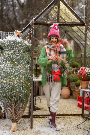 Téléchargez les photos : Jeune femme sort de belle serre décorée pour des vacances d'hiver à l'arrière-cour lors d'une chute de neige. Concept de bonnes fêtes de fin d'année - en image libre de droit