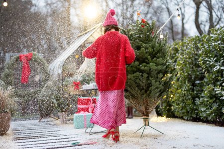 Foto de La mujer joven en suéter rojo y sombrero desempaca el árbol de Navidad de la red de embalaje, decorando el patio trasero para unas vacaciones de invierno. Concepto de preparación para un Año Nuevo y celebración - Imagen libre de derechos