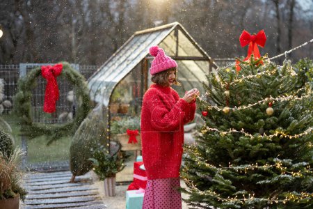 Téléchargez les photos : Une jeune femme en rouge décore un sapin de Noël luxuriant avec des balllets festifs et une guirlande dans la cour arrière de sa maison lors de chutes de neige, se préparant pour des vacances d'hiver - en image libre de droit