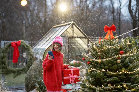 Téléchargez les photos : Femme en pull rouge et chapeau à l'aide d'un téléphone portable tout en se tenant avec une boîte-cadeau près de l'arbre de Noël à l'arrière-cour magnifiquement décorée lors d'une chute de neige. Concept de vacances d'hiver et de communication - en image libre de droit