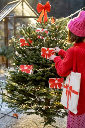 Téléchargez les photos : Femme décore arbre de Noël avec de grands arcs rouges à l'arrière-cour à l'extérieur. Concept de décoration pour des vacances d'hiver - en image libre de droit