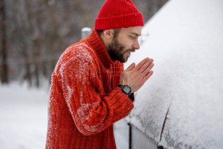 Téléchargez les photos : Man in red sweater and hat warms his hands while feeling cold at snowy backyard. Concept of winter time and cold weather - en image libre de droit