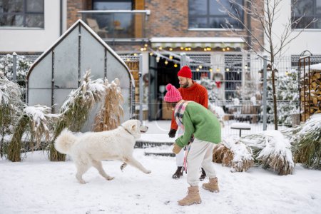 Téléchargez les photos : Young couple play with their adorable dog at snowy backyard, spending together winter time happily. Concept of winter holidays and leisure time - en image libre de droit