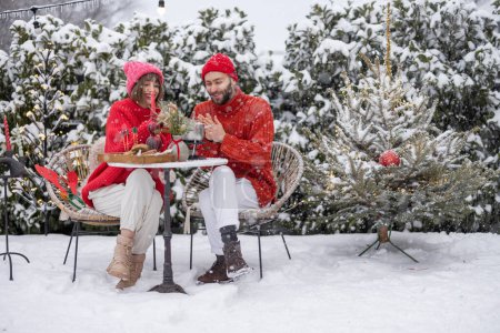 Téléchargez les photos : Man and woman have romantic dinner with fondue, while sitting together by the table at beautifully decorated snowy backyard. Young family celebrating winter holidays outdoors - en image libre de droit