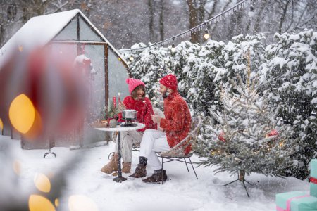 Photo for Man and woman have romantic dinner with fondue, while sitting together by the table at beautifully decorated snowy backyard. Young family celebrating winter holidays outdoors - Royalty Free Image
