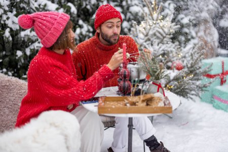 Téléchargez les photos : Man and woman in red winter clothes have fun while eating fondue, sitting together by the table outdoors at snowy garden. Young family celebrating winter holidays - en image libre de droit