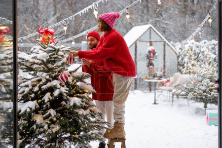 Téléchargez les photos : Man and woman decorate Christmas tree with festive balls, while preparing for a winter holidays at snowy backyard of their house. Happy family celebrating New Years holidays - en image libre de droit