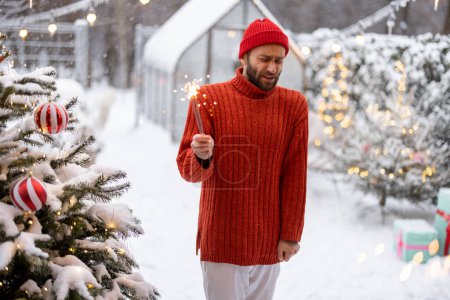 Foto de Hombre en suéter rojo celebra las vacaciones de Año Nuevo por la iluminación de brillantes cerca del árbol de Navidad en el patio trasero cubierto de nieve - Imagen libre de derechos