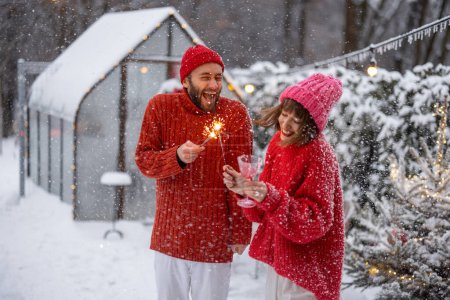 Photo for Man and woman in red sweaters celebrate New Years holidays by lighting sparklers and having fun near Christmas tree at snowy backyard - Royalty Free Image