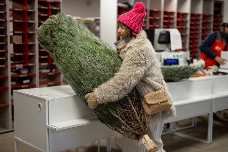 Photo for Young woman takes wrapped Christmas tree at delivery office. Concept of delivery service for the winter holidays. Happy woman picks up Christmas tree at post office - Royalty Free Image