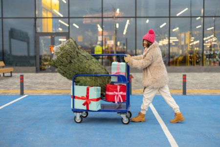 Photo for Woman pushing shopping trolley full of presents and Christmas tree in front of a mall outdoors. Winter holidays shopping concept - Royalty Free Image