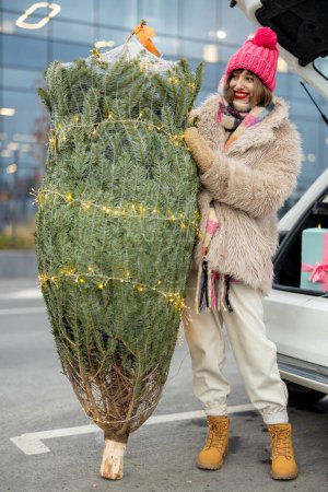 Téléchargez les photos : Jeune femme gaie se tient avec arbre de Noël enveloppé décoré avec guirlande près du coffre de voiture plein de cadeaux, ayant des vacances d'hiver shopping au centre commercial - en image libre de droit
