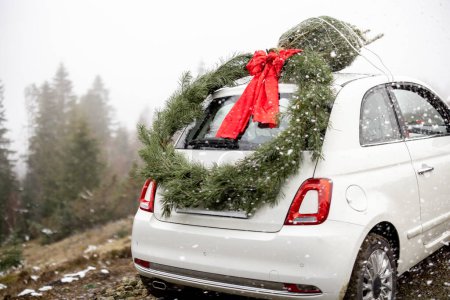 Téléchargez les photos : Voiture blanche décorée d'une grande couronne de Noël sur la route de montagne par temps neigeux. Concept du Nouvel An humeur et préparation pour les vacances - en image libre de droit